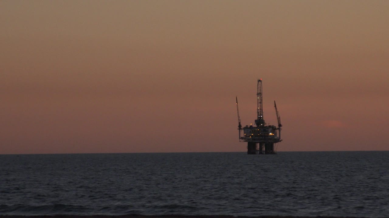Offshore oil platform near the center of the Huntington Beach frame, silhouetted over choppy dark water beneath a warm sunset sky with no visible people.