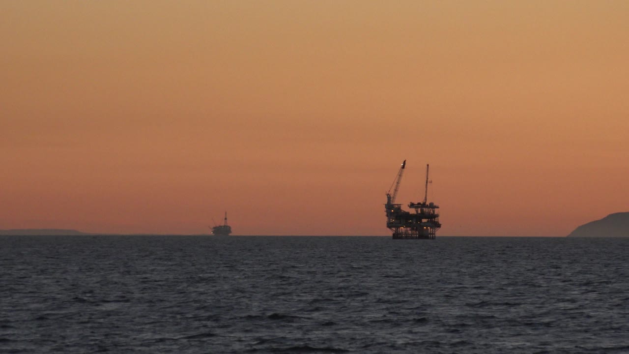 Huntington Beach ocean horizon at sunset with the main platform slightly right of center, a small vessel farther back, low background landform, textured water, and no visible people.