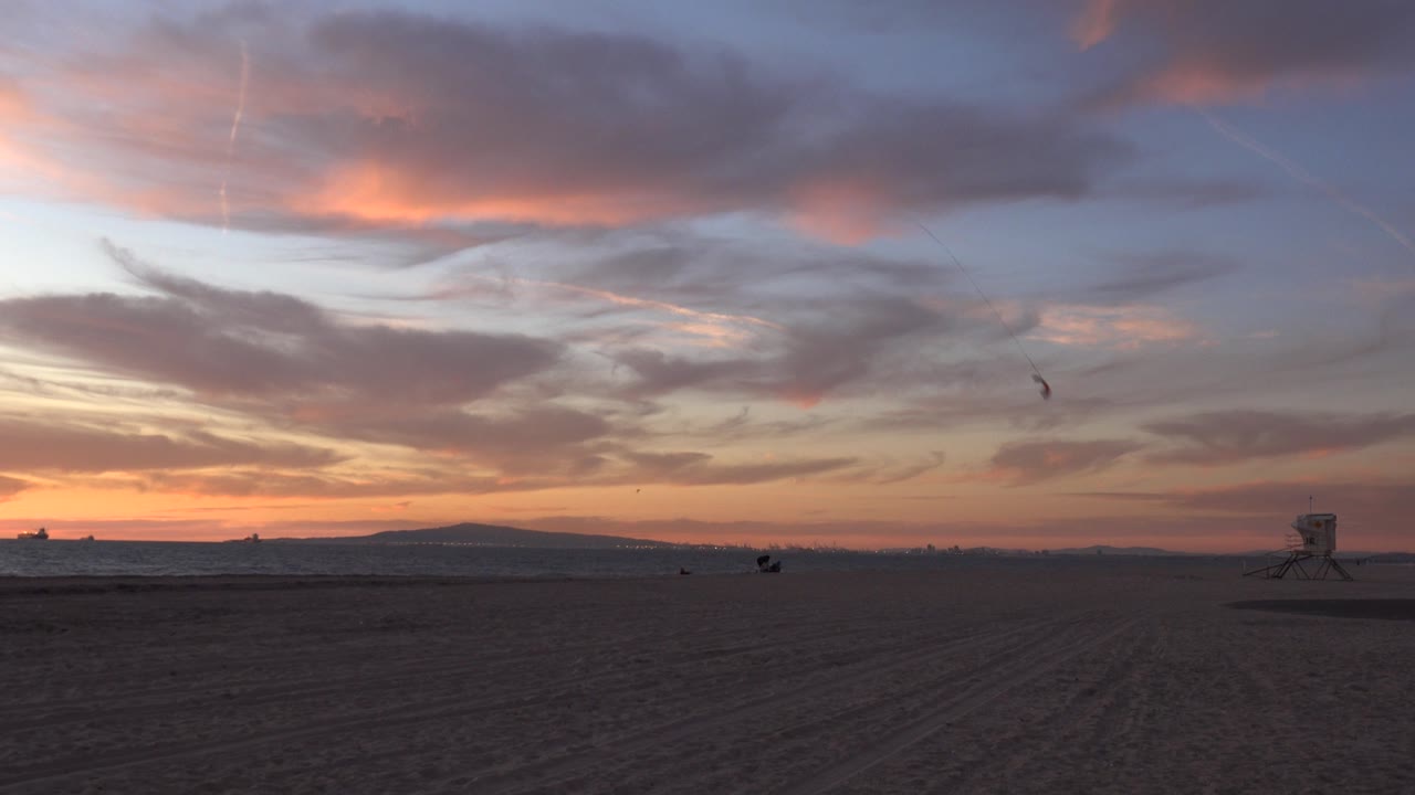 Wide view of Huntington Beach at sunset with dark sand in the foreground, ocean on the horizon, a lifeguard tower at right, and a small kite high in the clouded sky.