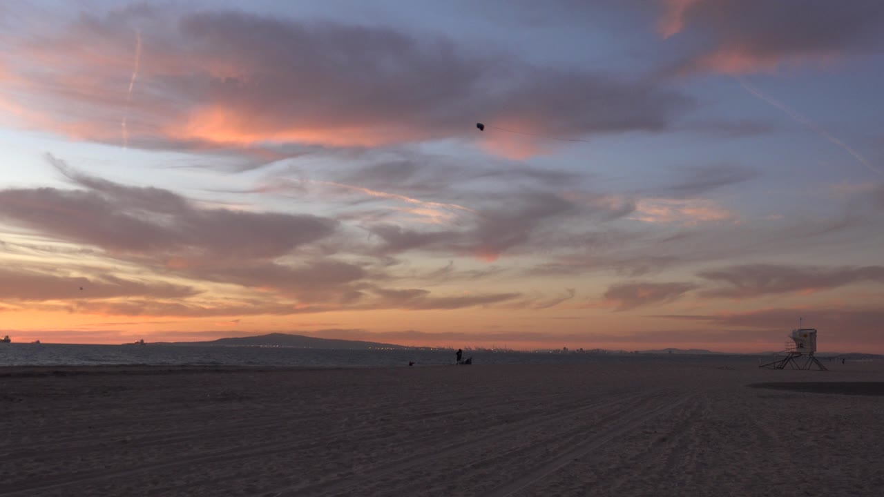 Opening frame of Huntington Beach, California, with orange cloud bands above a dark sandy foreground, the ocean horizon, distant low hills, a small lifeguard tower at right, and no visible people.