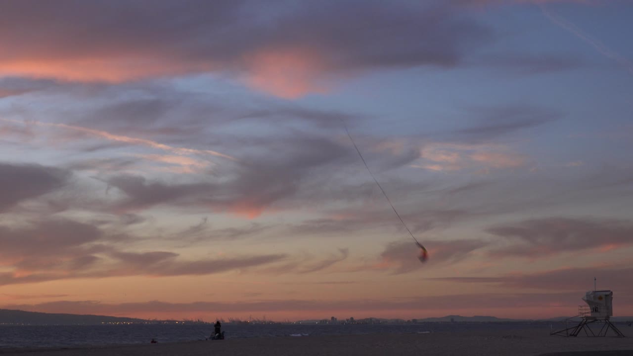 Huntington Beach dusk frame with layered pink and blue-gray clouds, a dark shoreline, a small airborne kite near center-right, a lifeguard tower at right, and distant tiny people visible near the waterline.