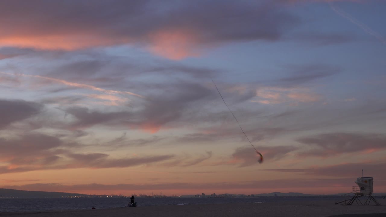 Huntington Beach sunset view with the kite or canopy slightly lower in the right sky, the lifeguard tower near the right edge, low silhouetted beach figures, and distant landforms along the horizon.