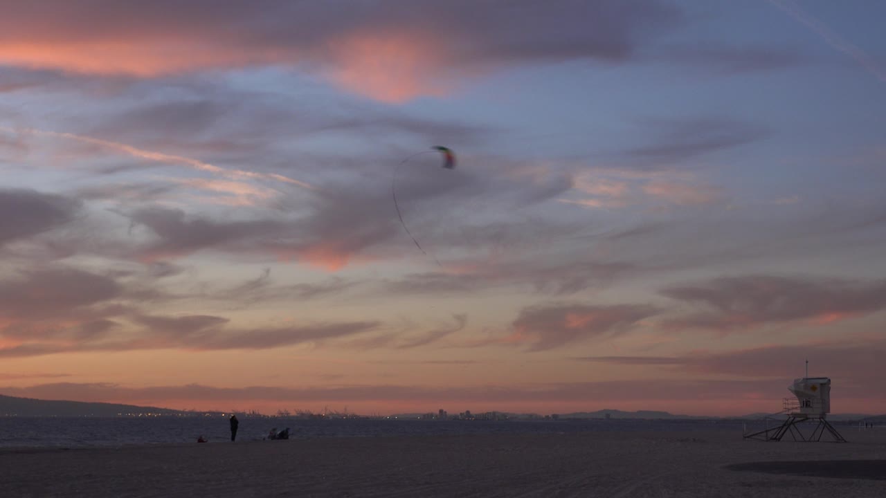 Huntington Beach shoreline at dusk with the kite slightly farther across the sky, small people on the sand, soft surf, and the lifeguard tower still visible to the right.