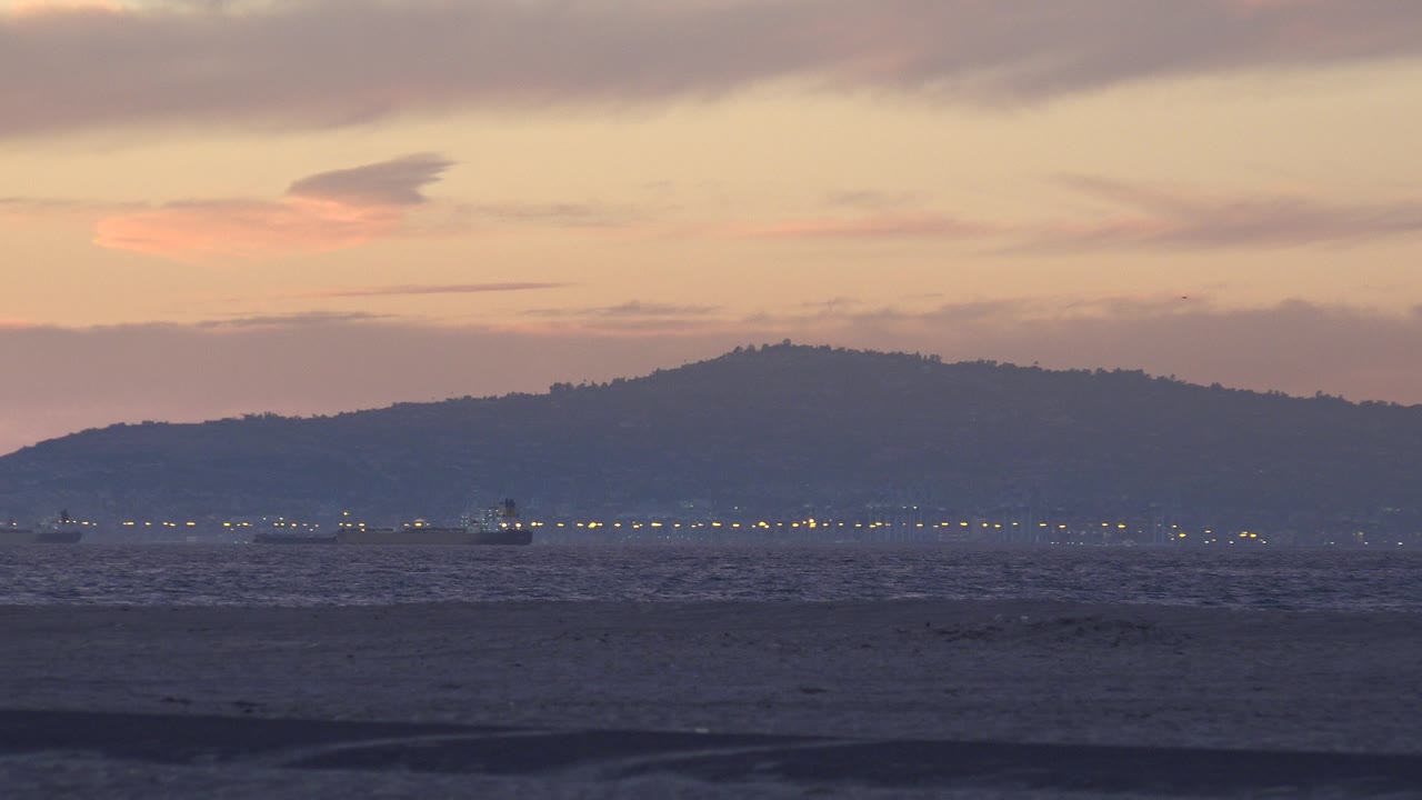 Huntington Beach, California dusk view with dark surf in the foreground, small shoreline lights on the horizon, distant hill silhouettes, peach clouds, and no visible people.