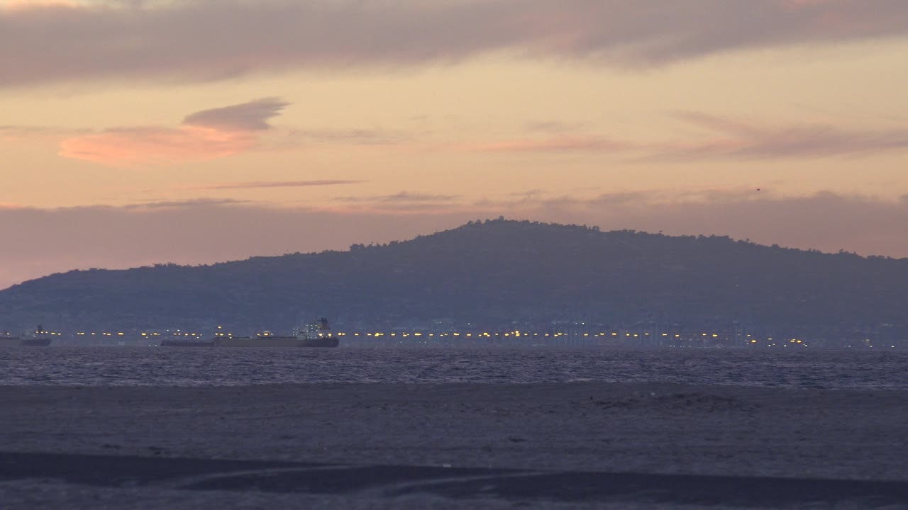 Huntington Beach, California sunset frame with low dark waves, a thin row of lights near the horizon, blue hills in the distance, soft peach sky, and no visible people.