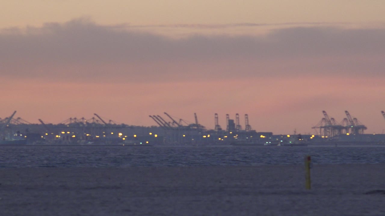 Huntington Beach sunset view with dark shoreline foreground, distant port cranes on the horizon, pink clouds overhead, and no visible people.