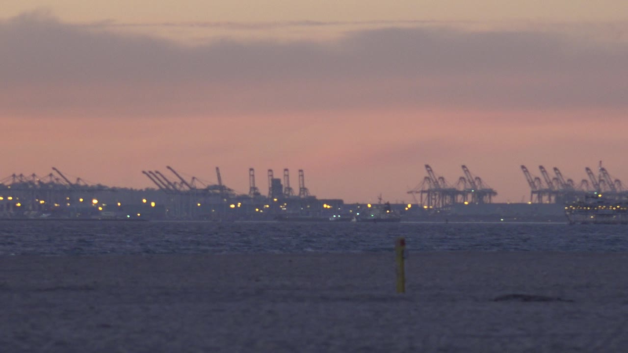 Huntington Beach dusk view with dark sand in the foreground, distant harbor cranes on the horizon, scattered lights, and no visible people.