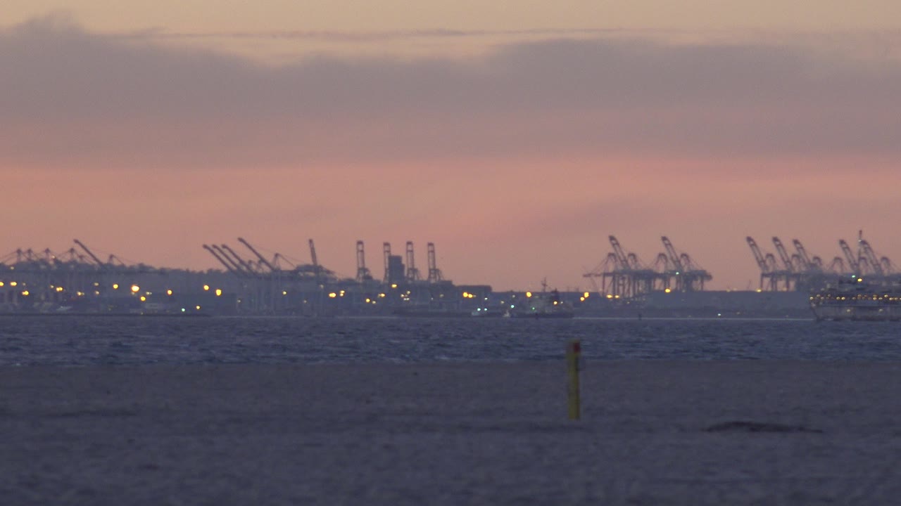 Huntington Beach shoreline at dusk with a low dark beach, faint surf texture, distant port cranes, small yellow lights, and no visible people.