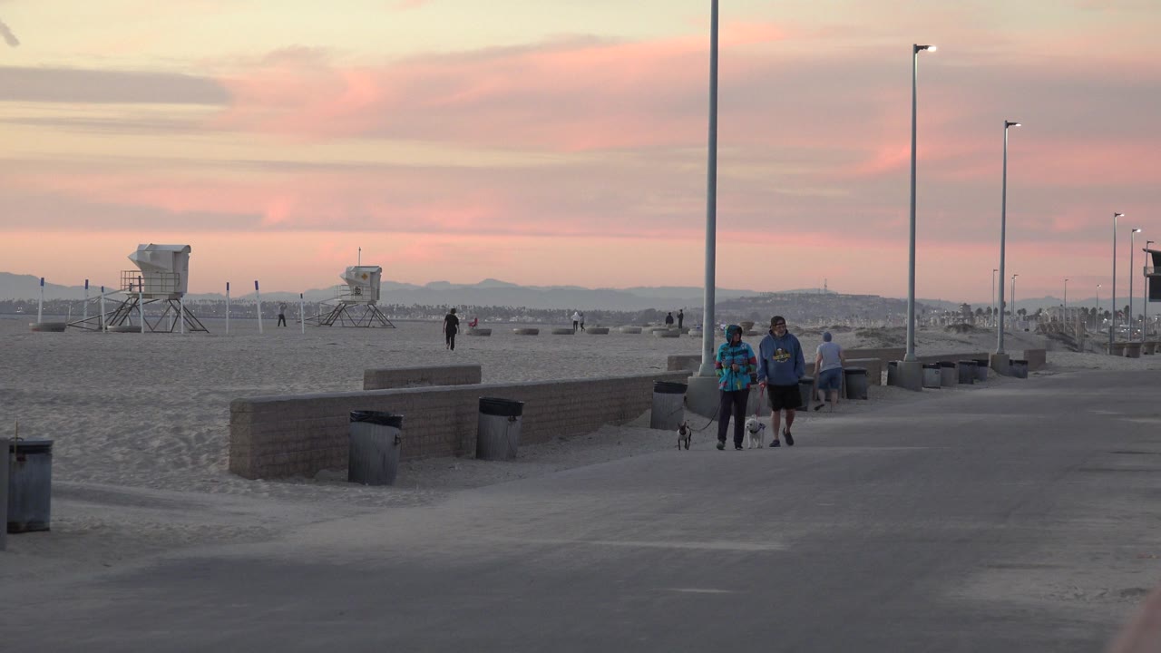 The two adults and small dog remain near the center of the Huntington Beach walkway, with a lifeguard tower and low beach walls behind them at sunset.