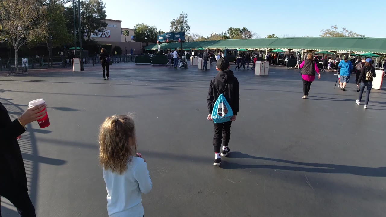 At Disneyland California, children and adults walk across a broad paved area toward green entrance canopies, with a child in a white top in the foreground and visitors ahead.