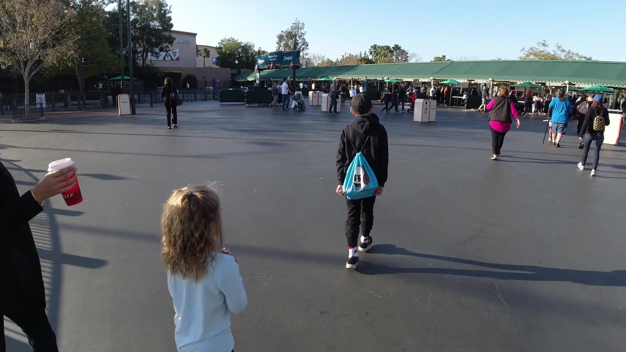 At Disneyland California, the walking group remains centered on the paved approach, with a cropped adult holding a red cup at left and families moving toward the green gate structure.