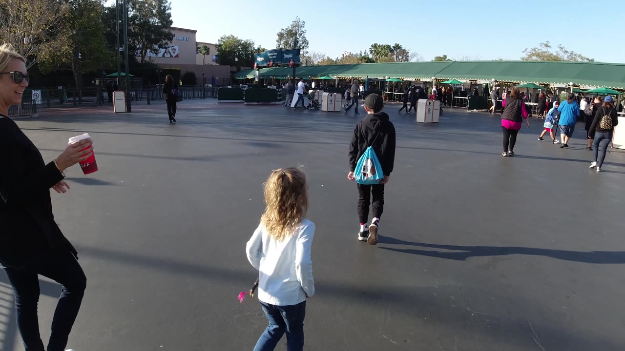 At Disneyland California, a blonde woman holding a red cup crosses the foreground beside a child in white, while other visitors walk toward the green entrance canopy.