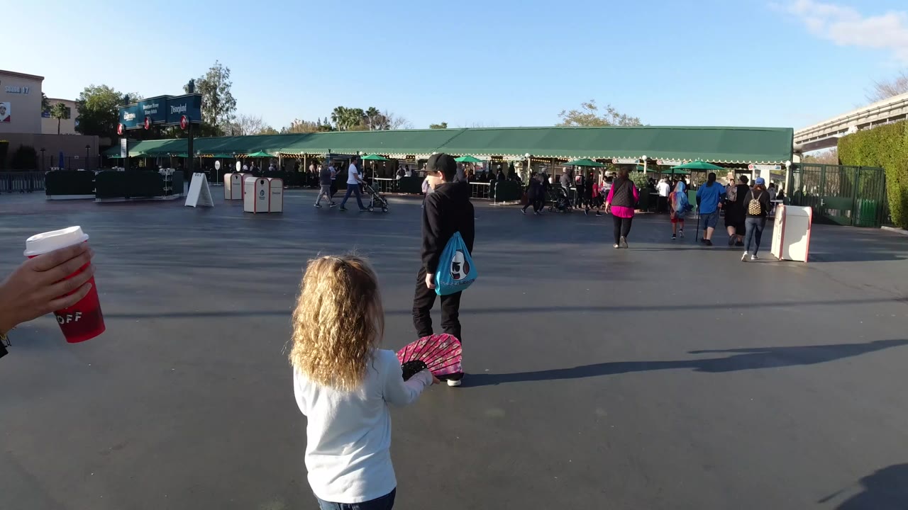 At Disneyland California, a young girl with curly light hair stands in the foreground while guests walk toward green awnings across a wide paved plaza.