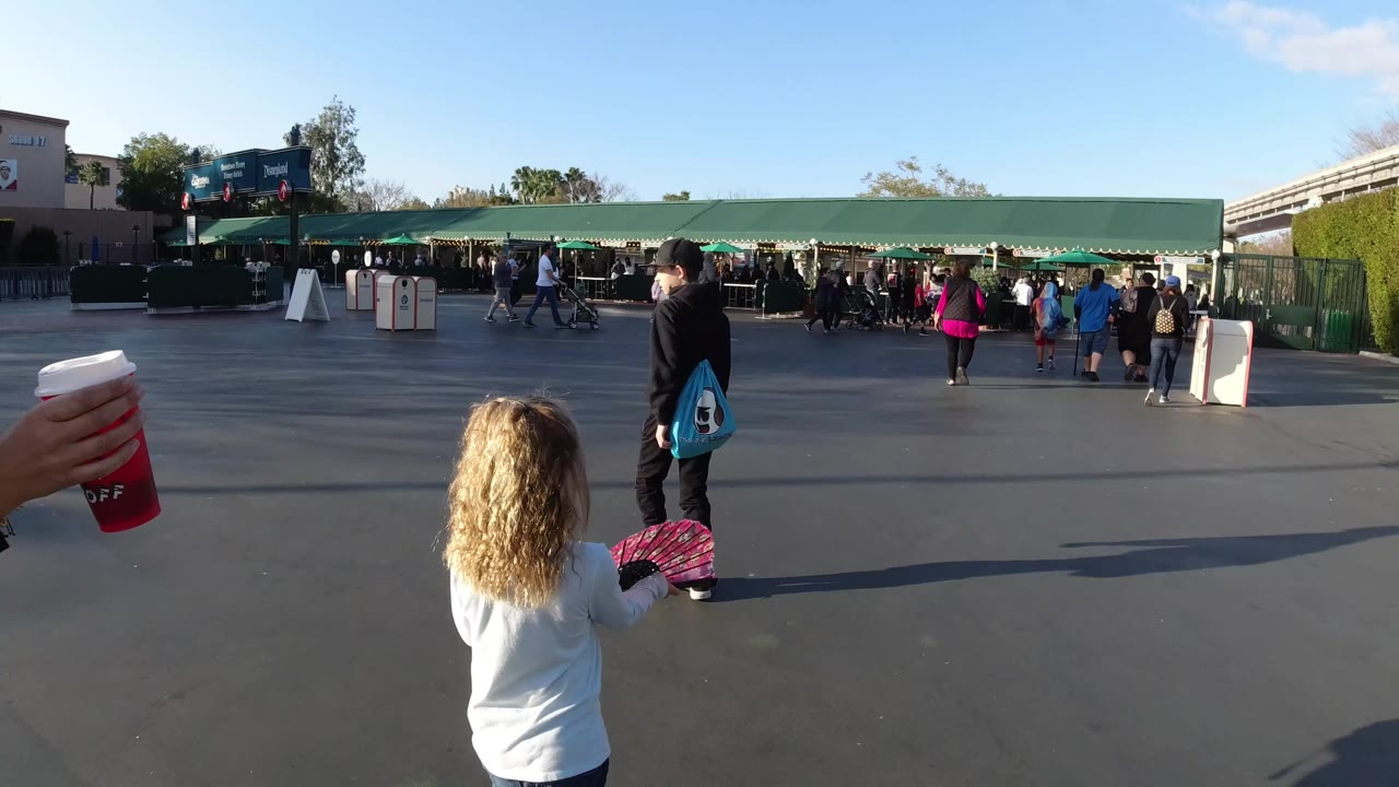 At Disneyland California, the girl remains near the foreground as a boy in black clothing stands farther right and visitor lines gather under green awnings.