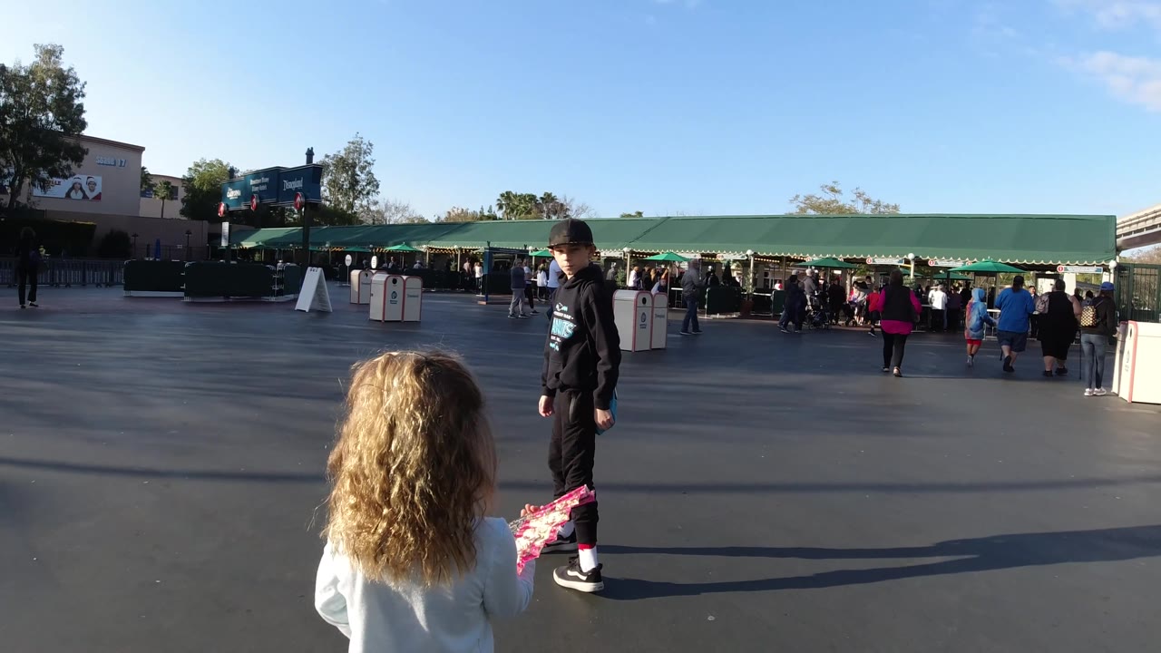 At Disneyland California, a boy in a black hoodie faces the camera near a younger girl, with green awnings, guests, and a hedge-lined structure in the background.