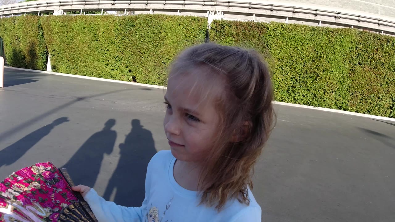 At Disneyland California, the girl looks off to the side while holding the floral fan higher in frame against the sunlit hedge and paved walkway.