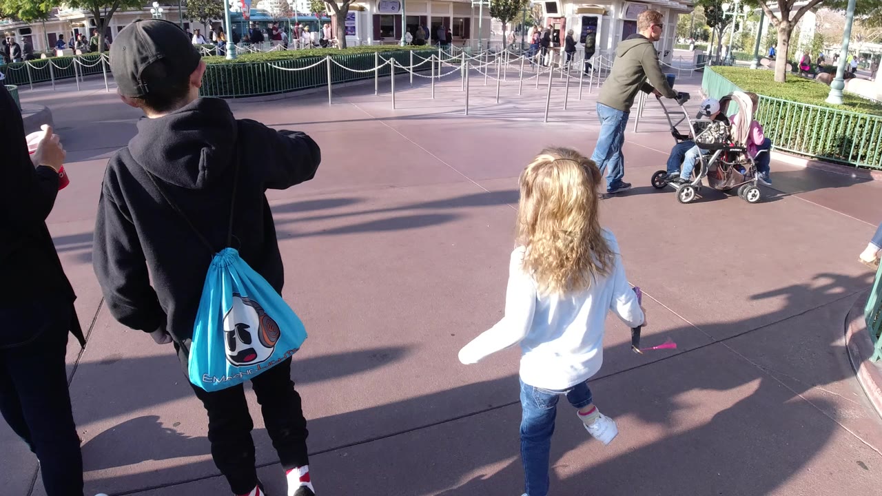 At Disneyland California, two children walk away from the camera on a reddish paved plaza while an adult pushes a stroller in the background and another hand holds a drink at the edge of frame.