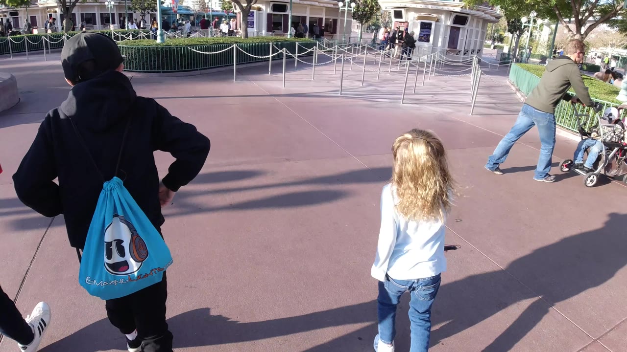 At Disneyland California, a boy in a black hoodie with a blue character bag and a girl in a white shirt walk away from the camera beside an adult's arm, with queue lanes and storefronts ahead.