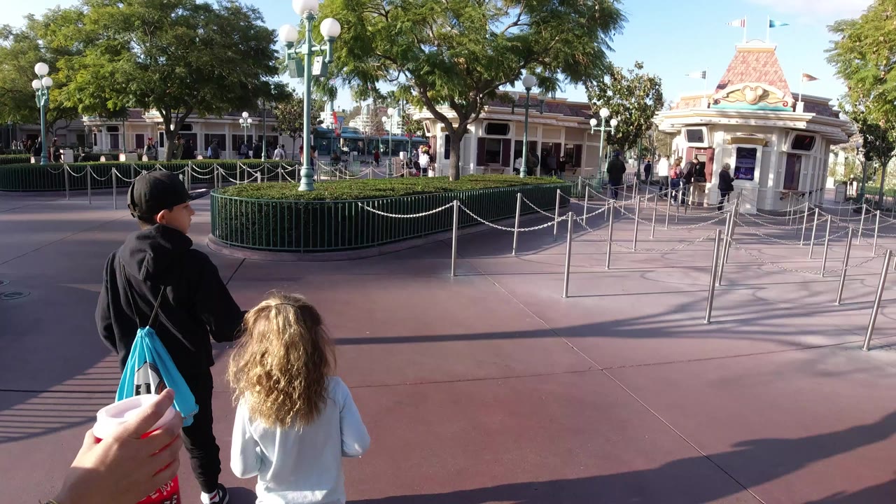 At Disneyland California, two children walk ahead from behind while an adult hand with a red drink cup enters the foreground near rope queues, hedges, lamps, and entrance buildings.