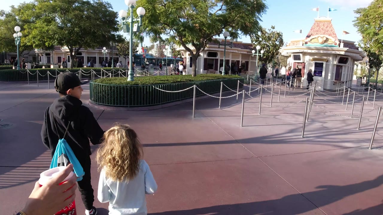 At Disneyland California, the boy and blonde child move farther along the pavement while the adult hand with a drink remains at the lower left and queue lanes fill the middle of the frame.