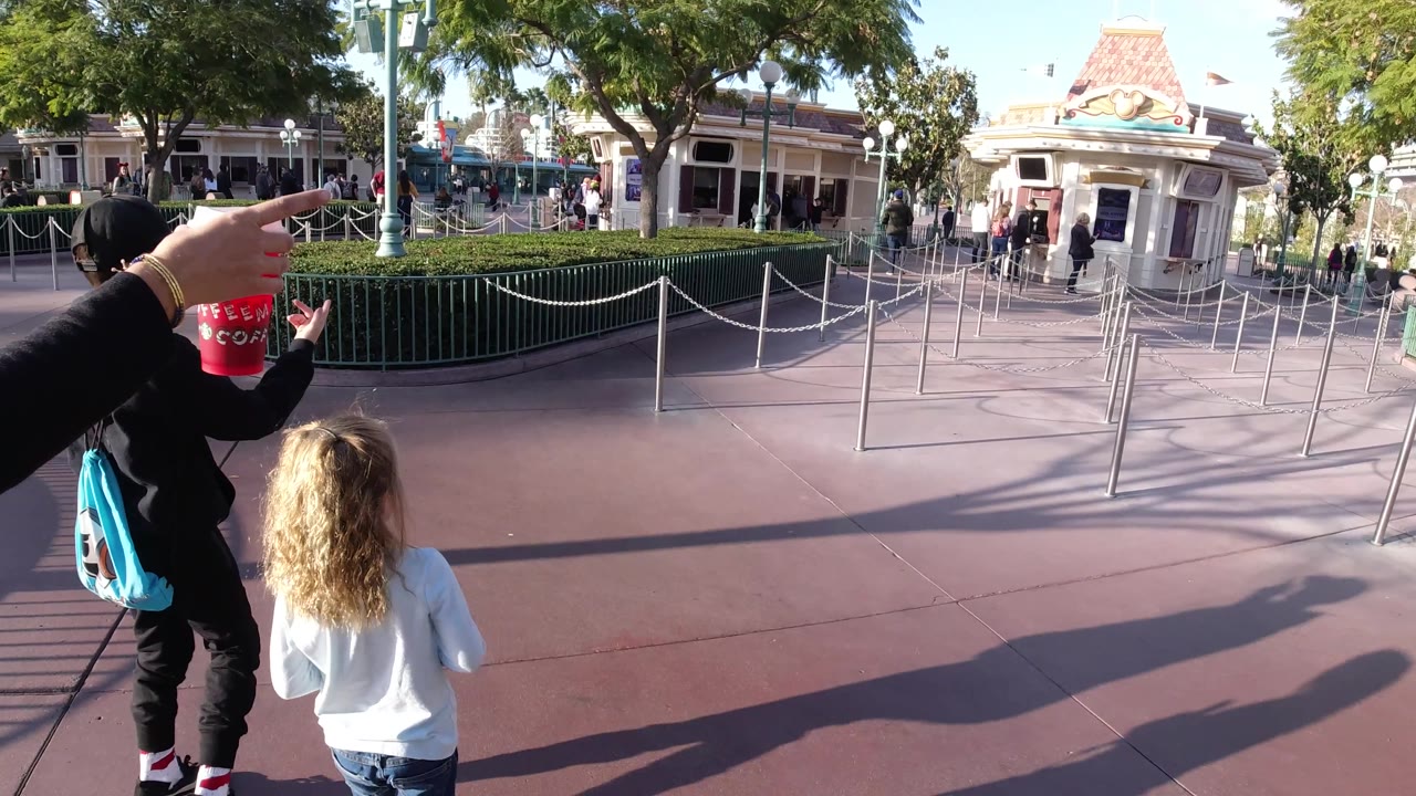 At Disneyland California, an adult arm points toward the right side of the walkway as the children pause or slow near the queue ropes, with hedges and a decorative park building behind them.