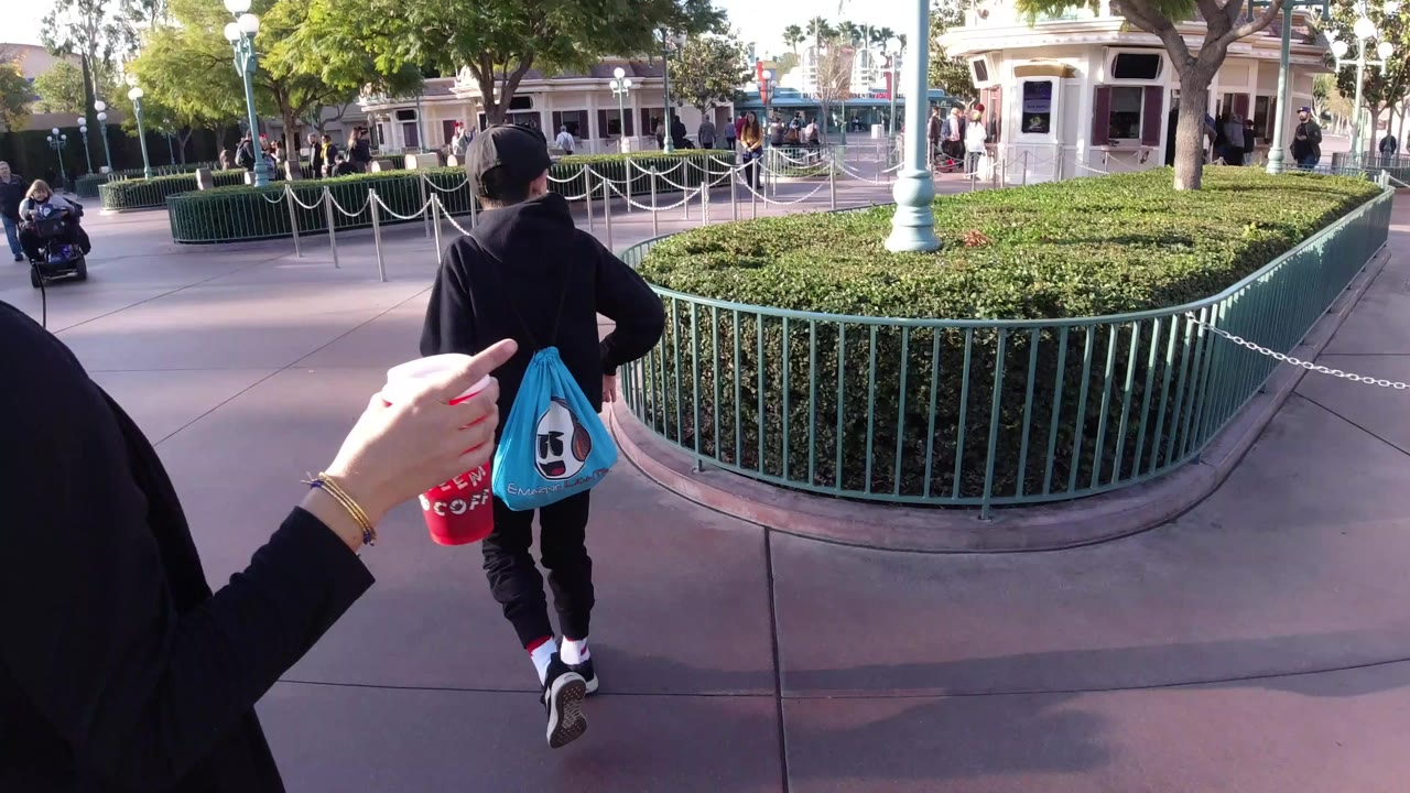 At Disneyland California, a foreground hand holding a red cup points toward a man in black walking beside green railings, hedges, and roped queue lanes.
