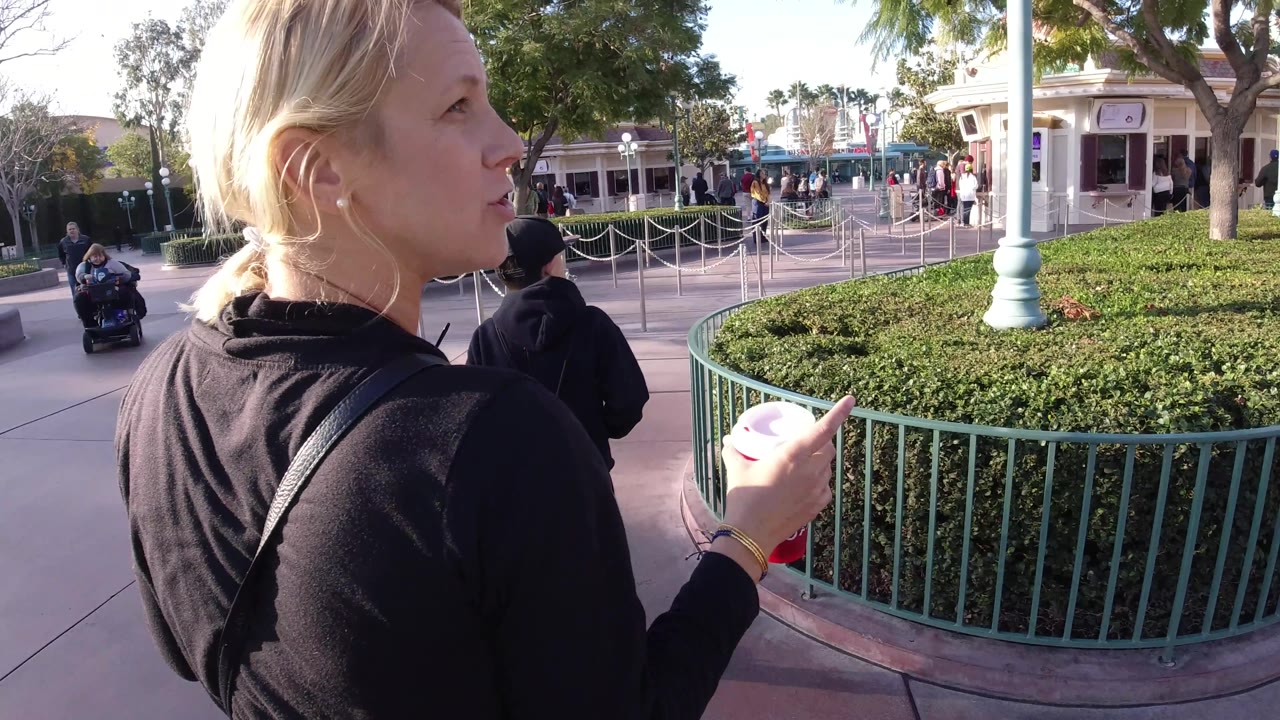 At Disneyland California, a blonde woman in a black sweater is seen close to camera in profile while holding a cup beside green railings, hedges, and queue lanes.