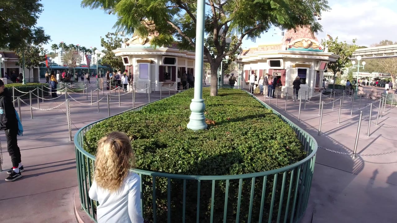 Disneyland California walkway view with a child seen from behind near green railings, trimmed shrubs, queue stanchions, trees, and visitors near a small themed building.