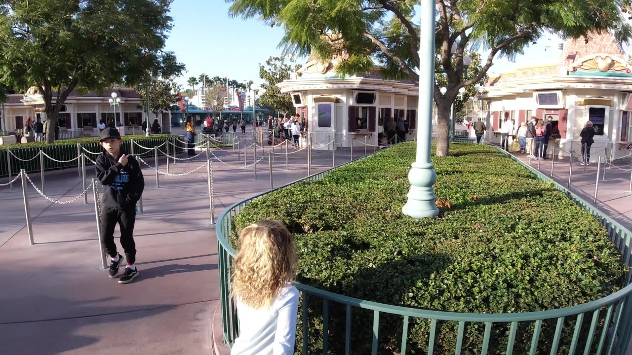 Disneyland California walkway frame with a boy near the right side, metal queue stanchions across the pavement, green shrub beds, trees, and visitors around the themed building.