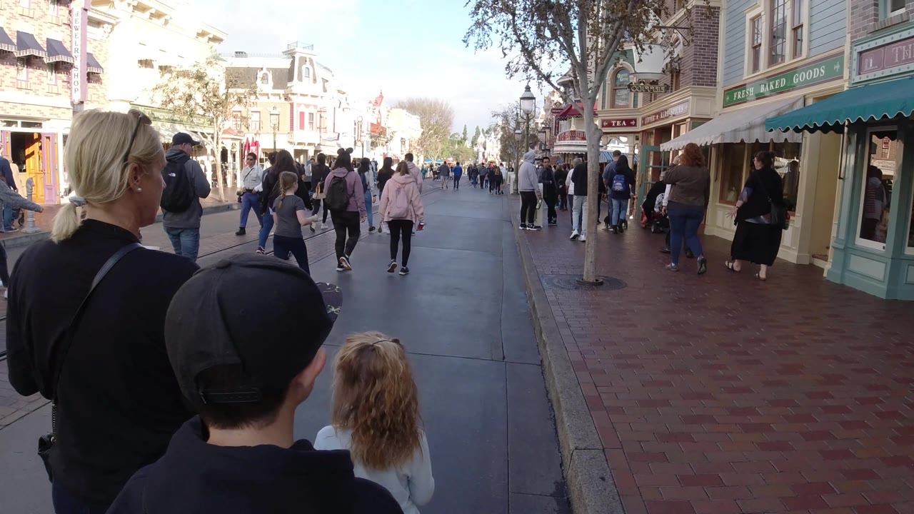 From a closer trailing angle, the woman and two children pass a storefront on the right while pedestrians and strollers fill the Disneyland California street ahead.