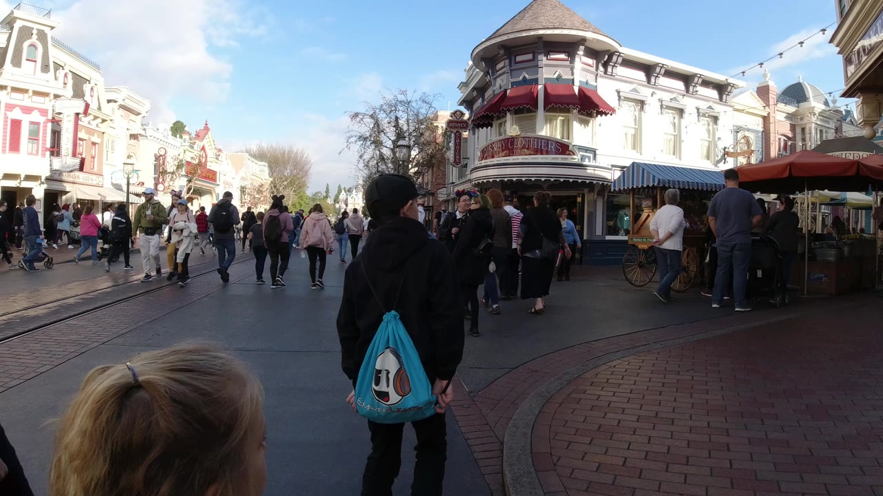 At Disneyland California, visitors move along a paved street lined with ornate storefronts, while a child's head and a person with a blue drawstring bag appear in the foreground.