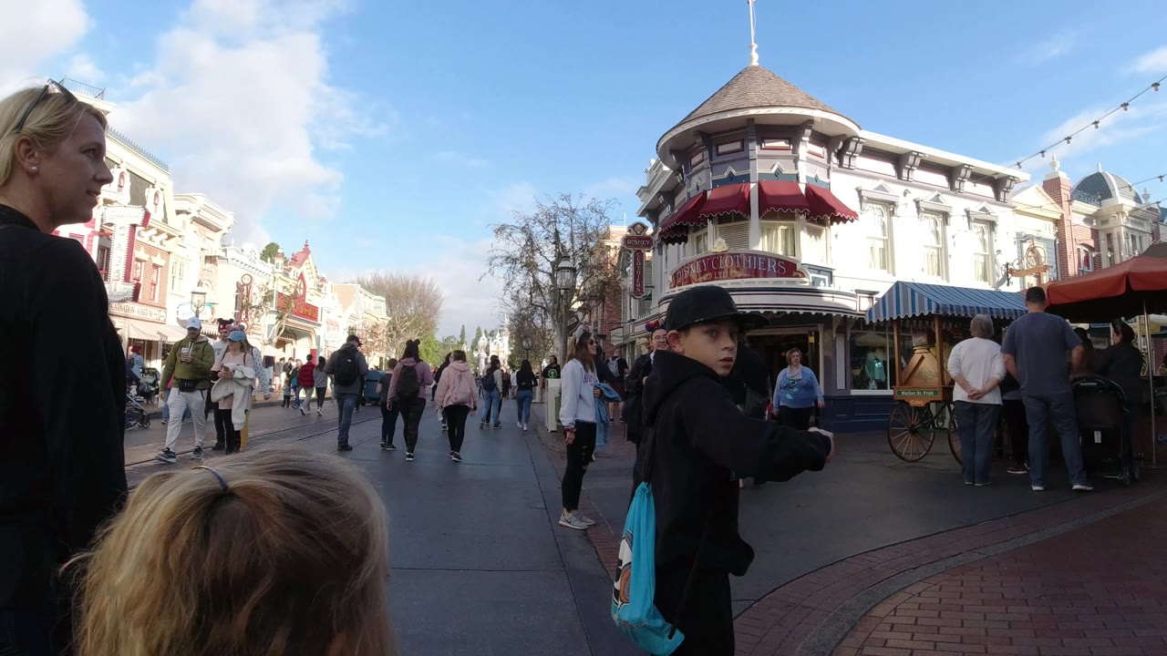 At Disneyland California, a woman in a black shirt stands close to the camera beside children, with visitors walking past themed storefronts in the background.