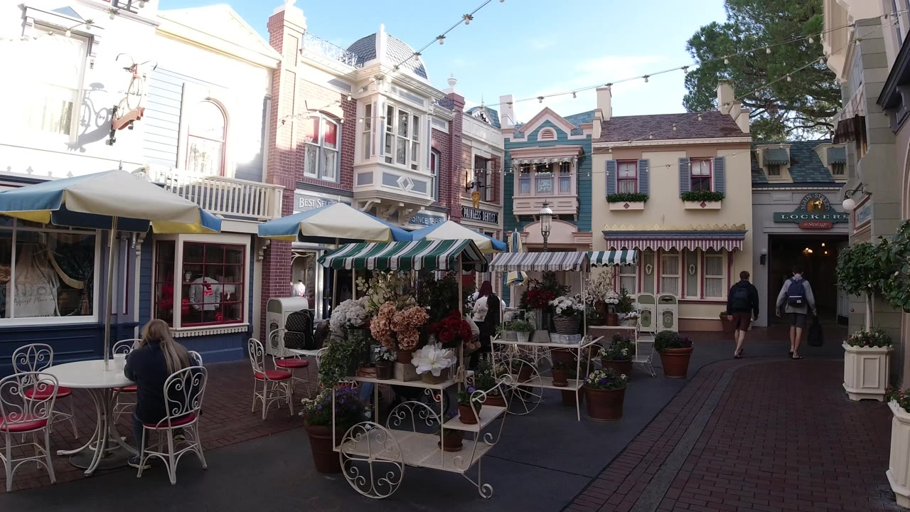 Disneyland California outdoor food court view with white cafe chairs, flower displays, striped awnings, pastel building facades, and people visible near the walkway.