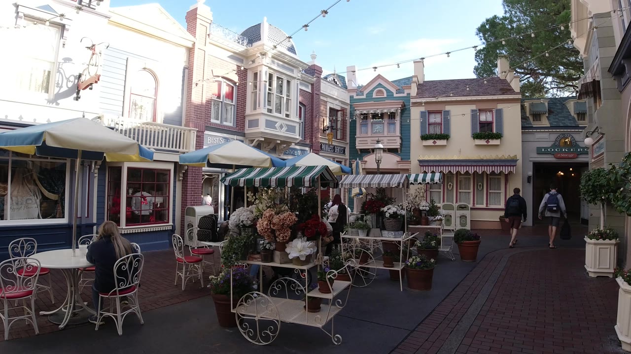 Disneyland California walkway scene with foreground cafe furniture, potted flowers, striped awnings, bright upper facades, and two people farther down the path.