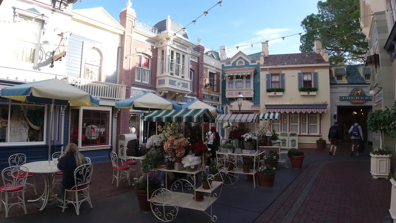 Disneyland California outdoor food court with a flower display centered in the foreground, cafe seating to the left, pastel storefronts behind it, and guests visible near the right-side entrance.