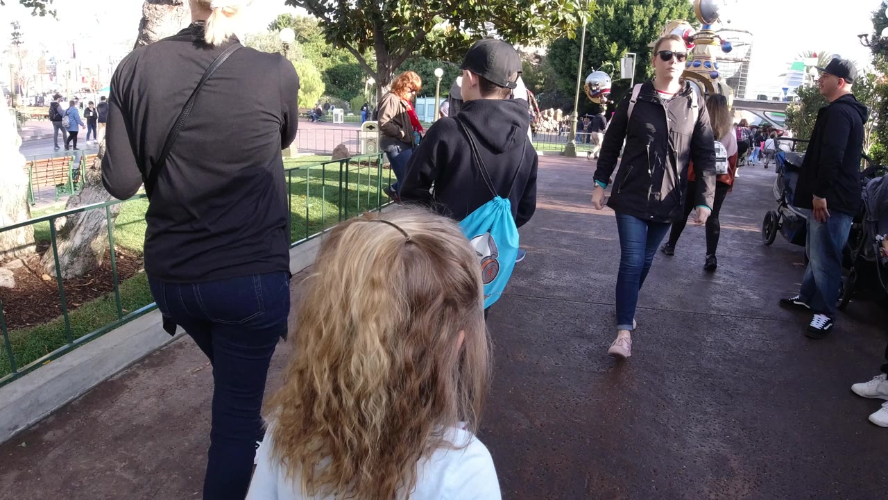 At Disneyland California, a pedestrian-height view follows visitors on a paved path, with a blond child in the foreground and people moving past railings and trees.