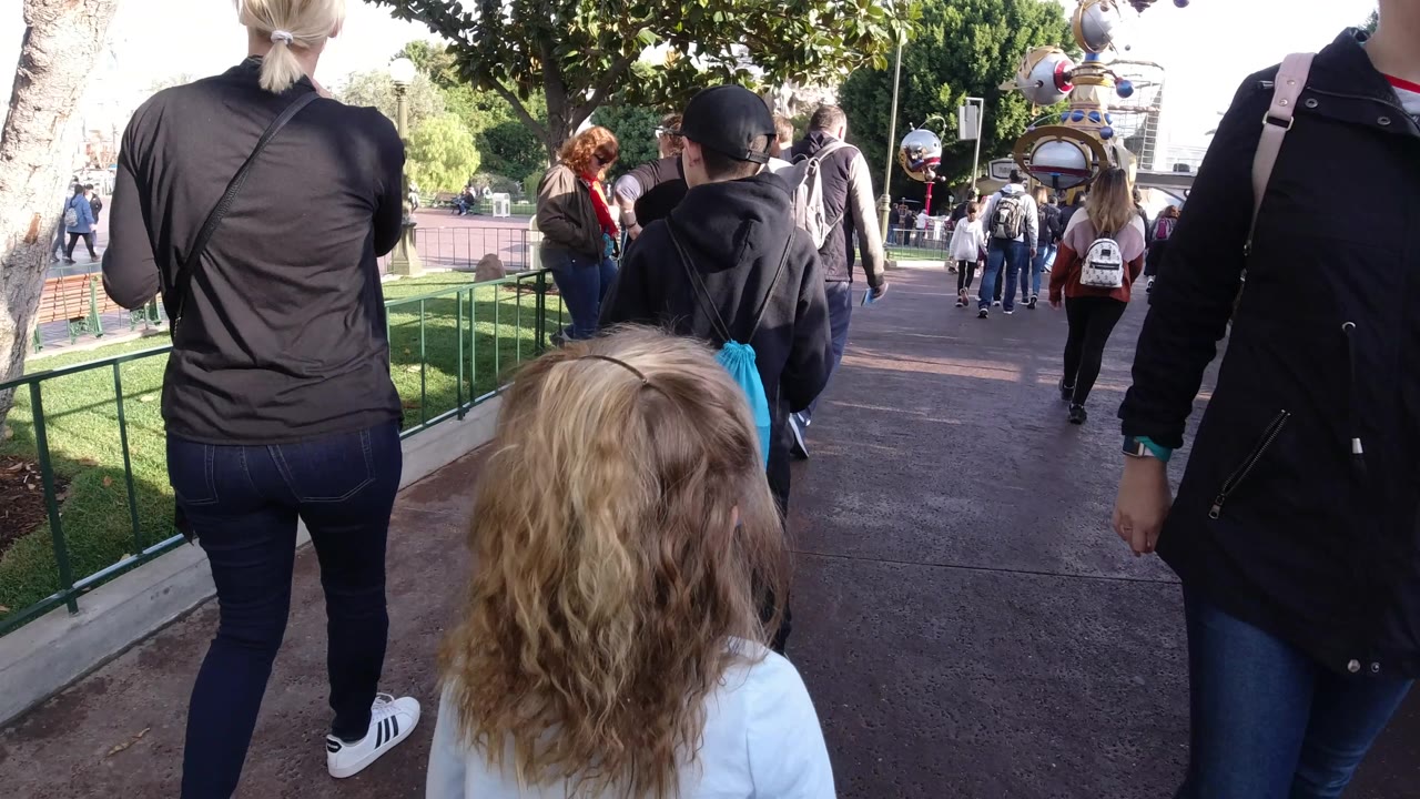 At Disneyland California, the viewpoint trails behind visitors, including a person with a bright blue drawstring bag, as the path opens toward more people and park structures.