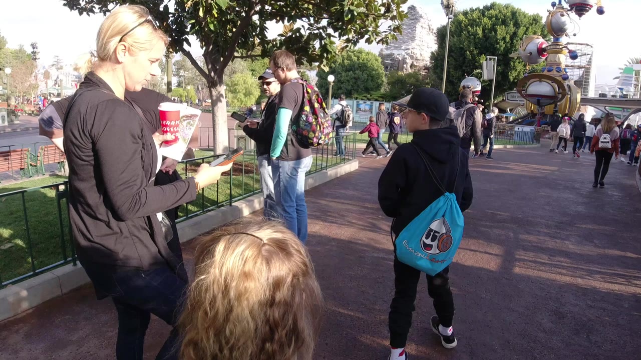 At Disneyland California, a woman in a black jacket holds a phone near two children as visitors move along a sunlit walkway with trees and railings behind them.