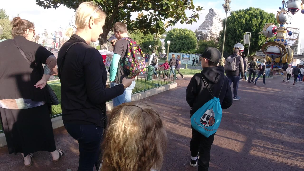 At Disneyland California, the group has shifted farther along the path, with the woman seen from behind and a child with a blue bag walking near green railings.