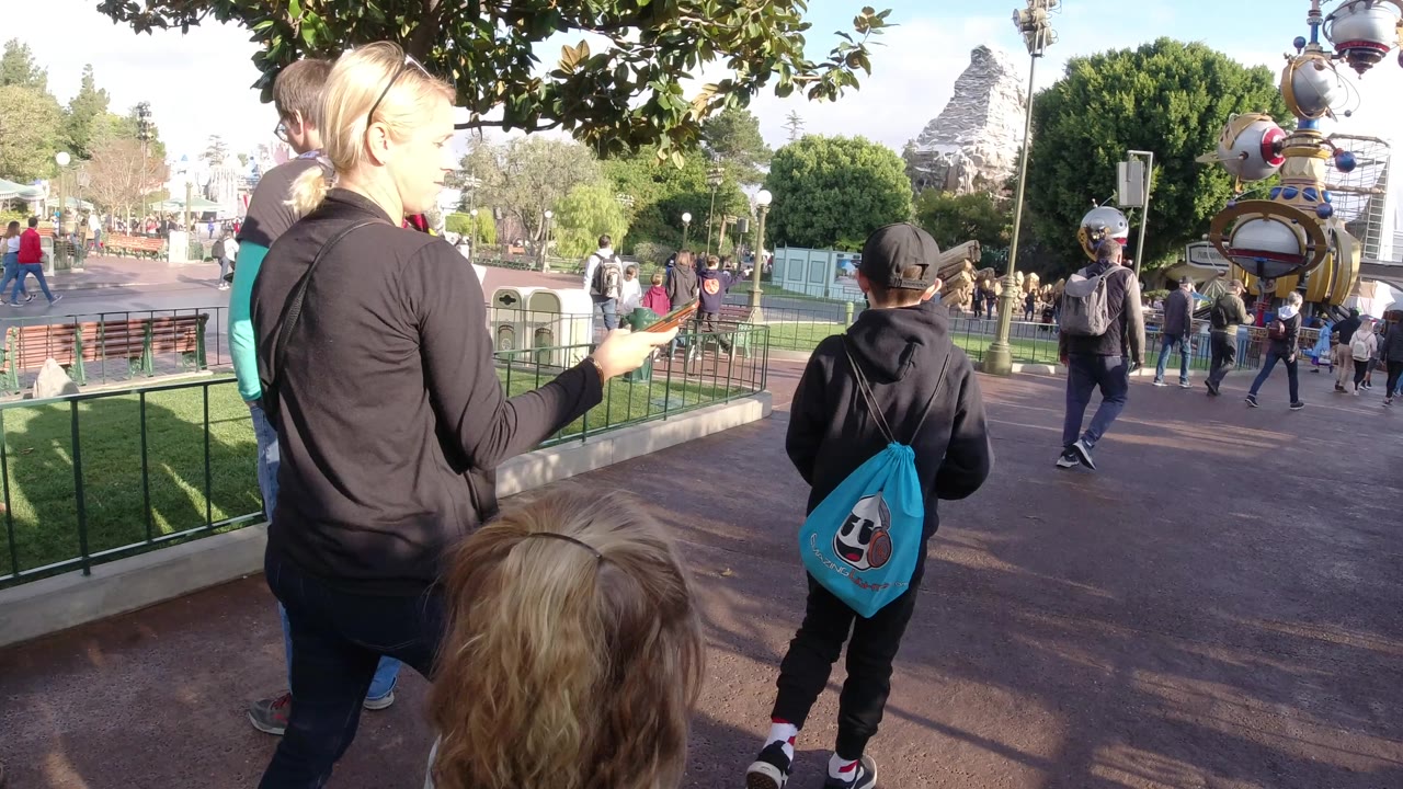 Woman in black jacket and children continue along the walkway under bright sun.