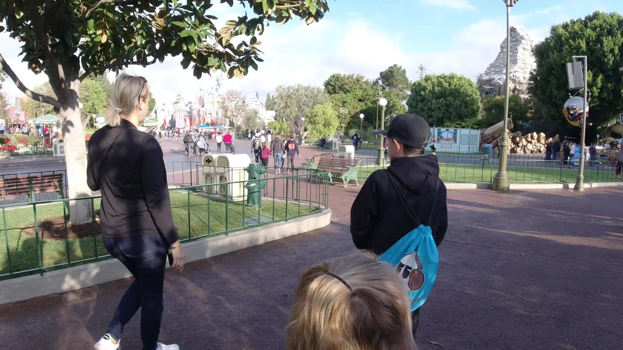 At Disneyland California, the walking group remains seen from behind, with the child carrying a bright blue drawstring bag beside an adult, railings and lawn to the right, and visitors farther down the walkway.