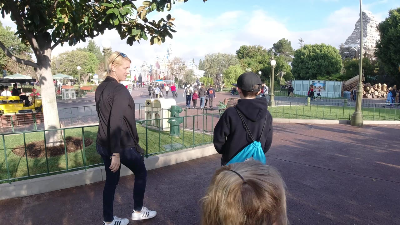 At Disneyland California, the adult turns partly toward the camera as the child in the black hoodie walks ahead with a blue bag, with a yellow park vehicle and crowded walkway visible in the background.