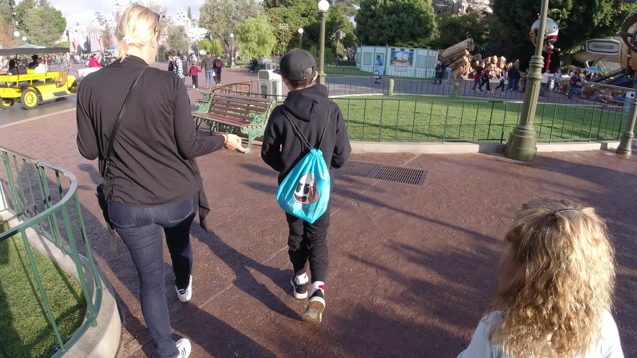 A woman in black and a boy with a bright blue drawstring bag walk away from the camera at Disneyland California, with a blonde child partly visible in the foreground and park benches, railings, trees, and visitors ahead.