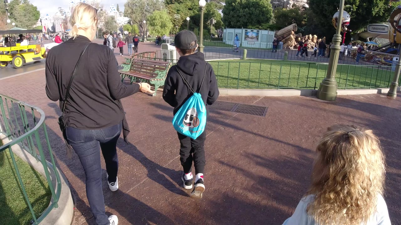 The woman and boy continue away from the camera at Disneyland California, with the blonde child at the lower edge, green railings on both sides, benches to the right, and a yellow park vehicle in the distance.