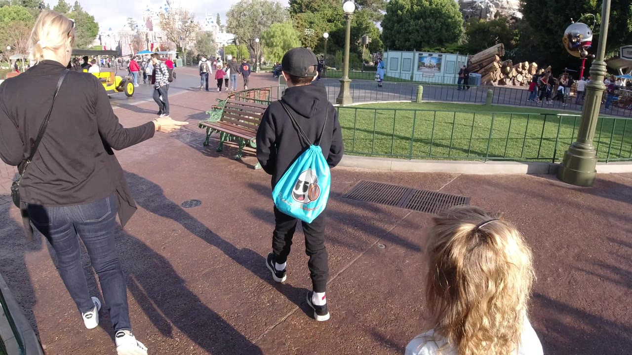 The woman raises one arm while walking with the boy at Disneyland California, as a blonde child remains low in frame and distant guests, benches, lampposts, trees, and a castle-like structure fill the background.