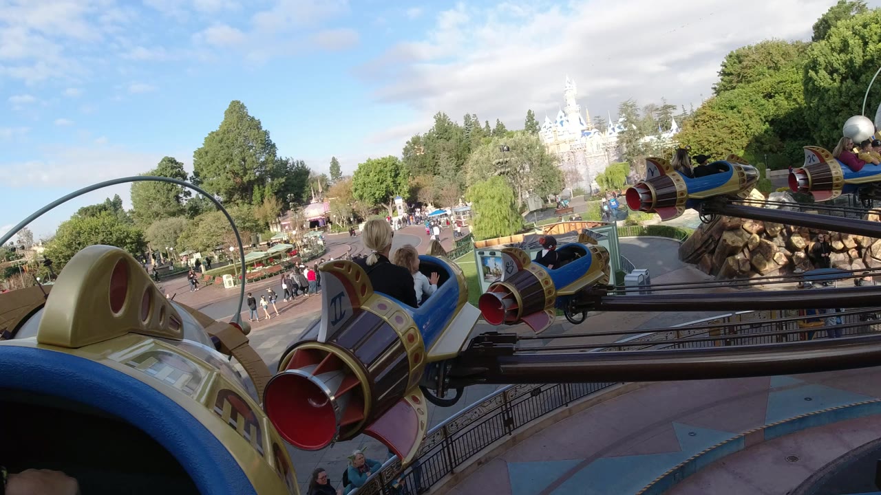 A Disneyland California ride view shows blue and gold rocket vehicles on metal arms, riders seated nearby, trees, walkways, and a pale castle-like structure in the distance.