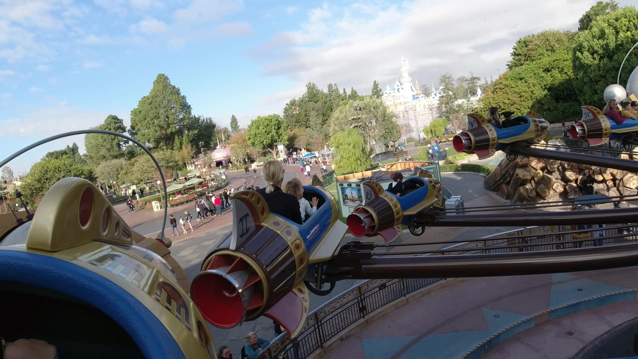 Nearby rocket ride cars pass over walkways and trees in bright daylight.