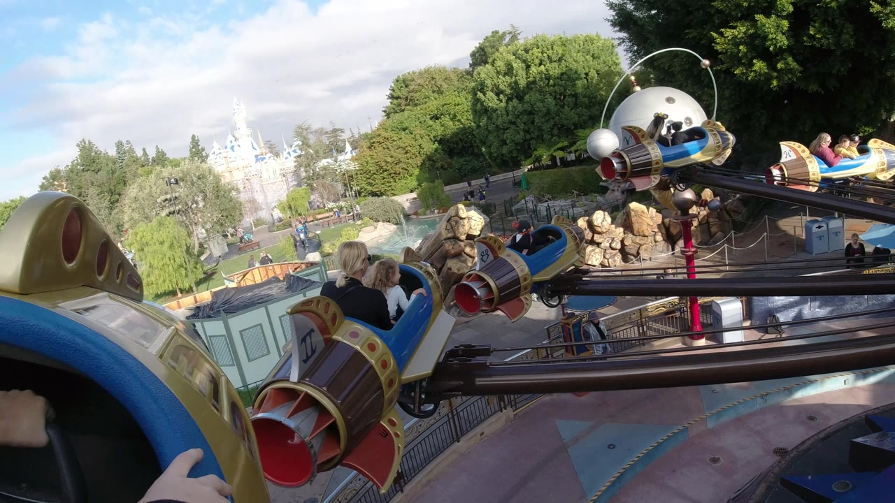 At 0:02, hands appear on the front edge of the ride vehicle as another Disneyland California rocket car passes close by with riders and park visitors visible below.