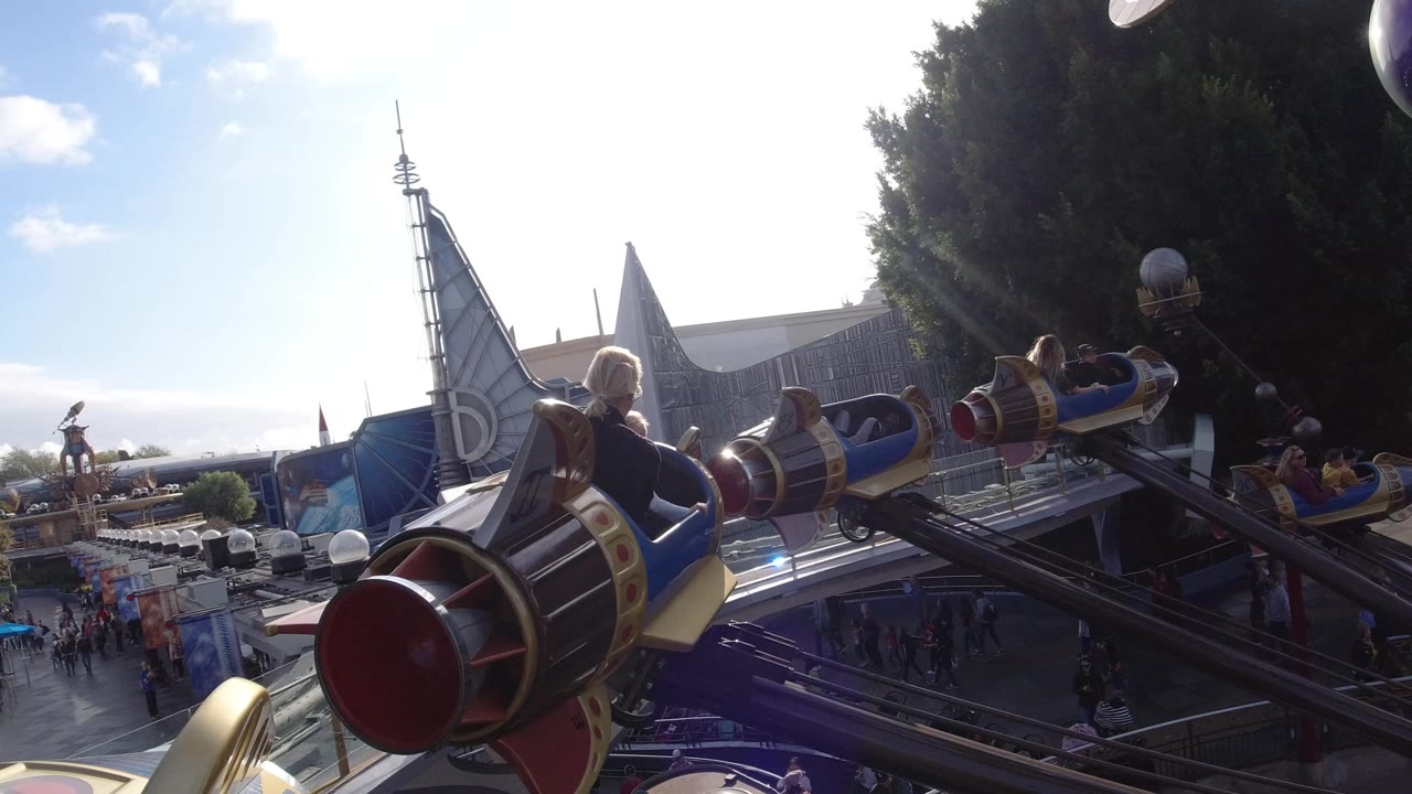 At Disneyland California, rocket-shaped ride cars extend over walkways and buildings in bright sun, with riders and park guests visible below.