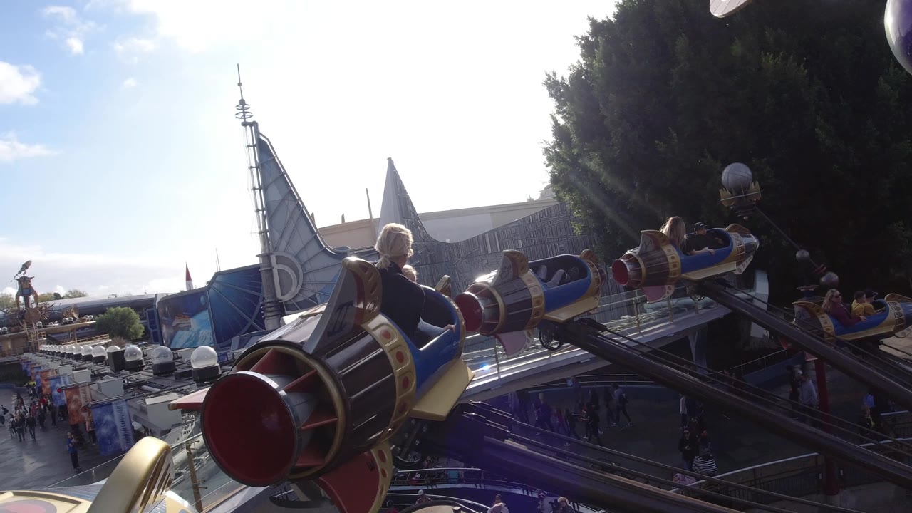 A close rocket-shaped vehicle with riders sits in the foreground at Disneyland California, with another car, a large tree, and park structures behind it.